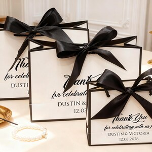 May include: White gift bags with black trim and large black satin bows. The bags have the text "Thank You for celebrating with us!" and names and a date printed on them. A pearl bracelet and gold tray are in the foreground.