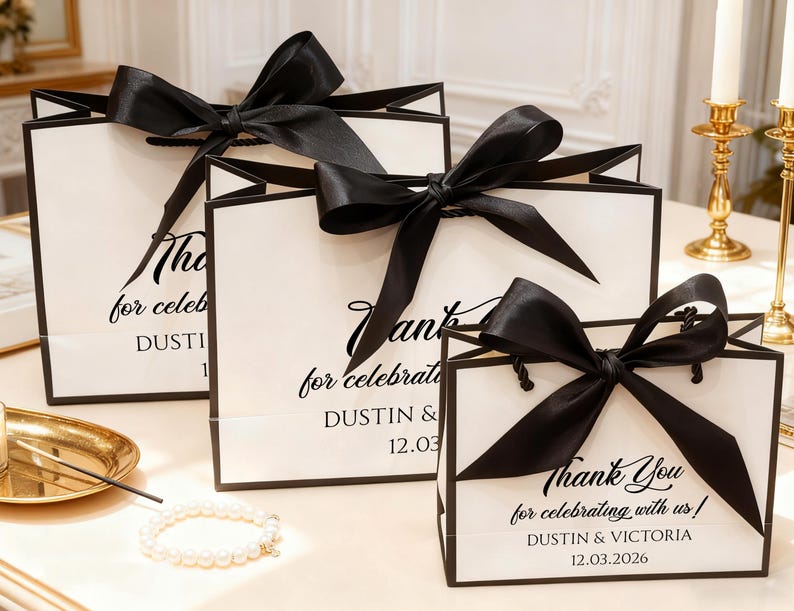 May include: White gift bags with black trim and large black satin bows. The bags have the text "Thank You for celebrating with us!" and names and a date printed on them. A pearl bracelet and gold tray are in the foreground.