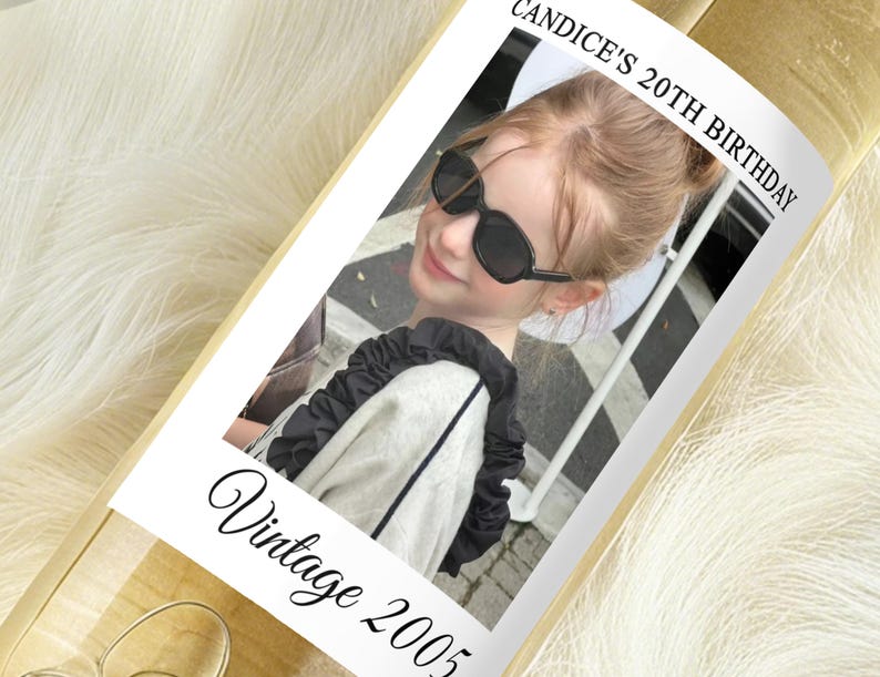 May include: A bottle with a label featuring a photo of a person wearing sunglasses. The label reads "Candice's 20th Birthday" and "Vintage 2005" in a script font. The bottle is on a white, fluffy surface.