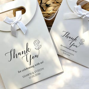 May include: Two white gift bags with a bow and the words "Thank You" in elegant script, along with a rose illustration. The text "for celebrating with us!" and names and a date are also printed on the bags. The bags are likely for a wedding or special event.