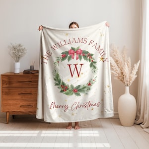 Può includere: Coperta natalizia color crema con un design a ghirlanda. La ghirlanda include agrifoglio, bacche e un fiocco rosso. Il testo recita "THE WILLIAMS FAMILY" e "Merry Christmas". Stelle dorate sono sparse sulla coperta.