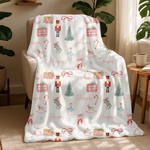 May include: White throw blanket featuring a festive pattern of Christmas-themed illustrations. The design includes nutcrackers, gift boxes, Christmas trees, ballet slippers, and the name "Harper". The blanket is draped over a beige armchair.