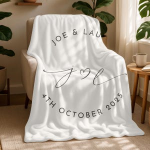 Op de afbeelding: Een witte fleece deken met zwarte tekst met de tekst "JOE & LAU", een gestileerde "j♡l" en "4TH OCTOBER 2025". De deken ligt over een stoel en is een gezellig woonaccessoire.