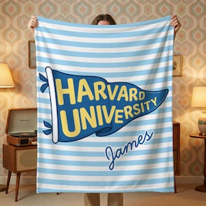 以下が含まれることがあります： 青と白のストライプのブランケット。紺色のペナントには黄色で「HARVARD UNIVERSITY」と書かれています。名前の「James」は筆記体で下に書かれています。ブランケットは人が持っています。家庭用アクセサリー。