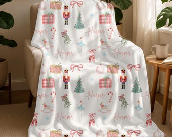 May include: A white fleece blanket featuring a repeating pattern of Christmas-themed illustrations including nutcrackers, ballet dancers, pointe shoes, gift boxes, candy canes, bows, and the name Harper written in a cursive pink script.