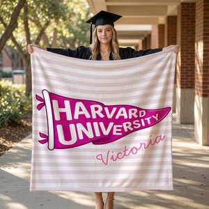 以下が含まれることがあります： ピンクと白のストライプのブランケット。フクシア色のハーバード大学のペナントデザインが特徴です。ペナントには筆記体で「Victoria」という名前が入っています。卒業キャップとガウンを着た人がブランケットを持っています。