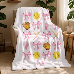 May include: White throw blanket featuring pink bows, softballs, baseball gloves, and drink tumblers. The blanket has a personalised name in pink script. The softball is yellow with red stitching. The baseball glove is brown and yellow.