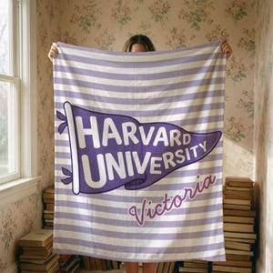 以下が含まれることがあります： ハーバード大学のロゴがペナントデザインで描かれた、紫と白のストライプのブランケット。ロゴの下には「Victoria」の文字が筆記体で書かれています。窓と本の山を背景にブランケットが掲げられています。