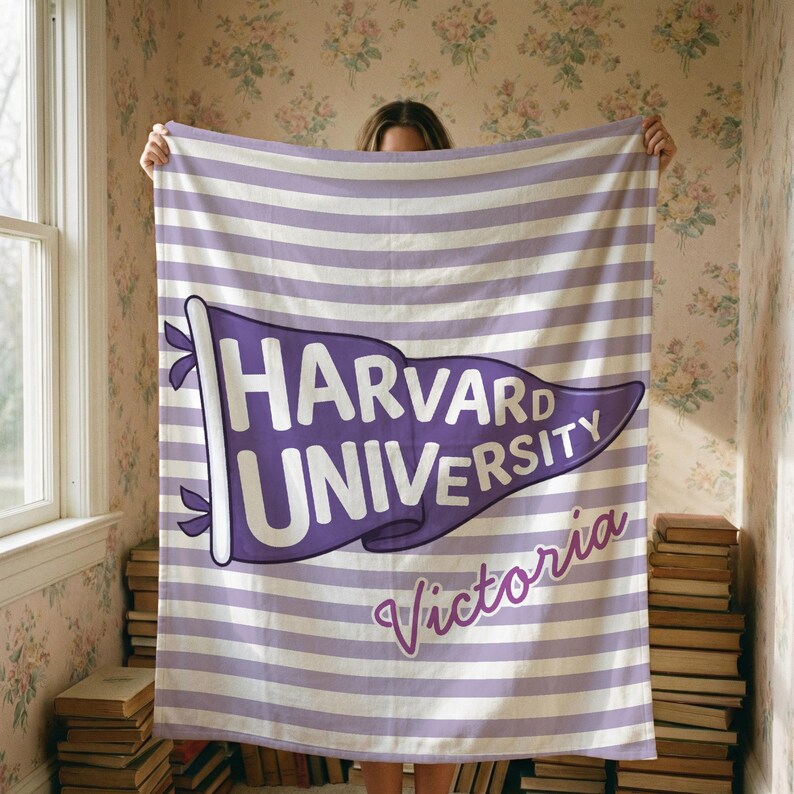 以下が含まれることがあります： ハーバード大学のロゴがペナントデザインで描かれた、紫と白のストライプのブランケット。ロゴの下には「Victoria」の文字が筆記体で書かれています。窓と本の山を背景にブランケットが掲げられています。