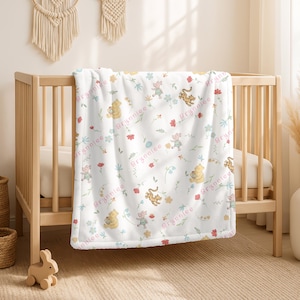 May include: A white baby blanket draped over a wooden crib. The blanket features a repeating pattern of Winnie the Pooh characters, flowers, and the name "Brynniee" in pink script. The blanket is soft and cozy.
