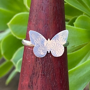 May include: A silver butterfly ring with a detailed design. The ring is on a wooden display stand.