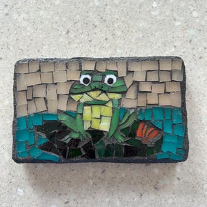 Peut inclure: Mosaïque représentant une grenouille verte et jaune sur un fond d'eau turquoise et de carreaux beiges. Le motif est encadré par une bordure sombre, créant une pièce décorative.