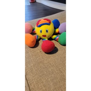 May include: A colorful plush octopus toy with a yellow head, blue eyes, and a red nose. It has a rainbow-striped body and eight tentacles in various colors, including pink, orange, green, and purple. The octopus wears a hat with red, orange, and yellow stripes.