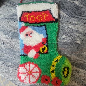 Puede incluir: Calcetín navideño hecho a mano con un diseño de Papá Noel. El calcetín es verde con una parte superior blanca y presenta una imagen de Papá Noel con la palabra "Toot" en rojo. El calcetín tiene una flor roja y blanca y una campana amarilla.