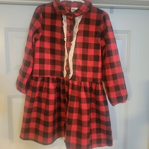 Puede incluir: Un vestido a cuadros rojos y negros con un ribete de encaje blanco en la parte delantera. El vestido tiene mangas largas y cintura fruncida. El vestido está colgado de una percha blanca.