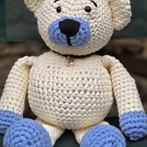 Peut inclure: Un ours en peluche crocheté en fil couleur crème avec des accents bleu clair sur les pattes et le museau. L'ours présente des yeux en plastique noir, un nez brodé noir et un collier foncé orné d'une petite perle et d'un minuscule cœur rose.