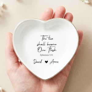 Può includere: Un piattino in ceramica bianca a forma di cuore con bordo dorato. Al centro è presente il testo nero The two shall become One Flesh, Ephesians 5:31, e i nomi Dand e Anna separati da un piccolo simbolo di cuore nero.