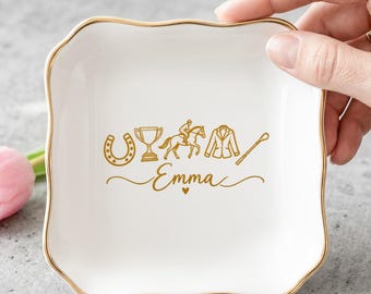 May include: A square white ceramic trinket dish with scalloped edges and a gold-colored rim. The center features gold-colored line art of a horseshoe, trophy, horse and rider, jacket, and crop, with the name Emma written below.
