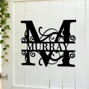 Könnte beinhalten: Schwarzes Metallschild mit dem Namen "Murray" innerhalb des Buchstabens "M". Das Schild hat dekorative Verzierungen und ist an einem weißen Holzzaun befestigt. Grüne Ranken und eine Laterne sind ebenfalls zu sehen.