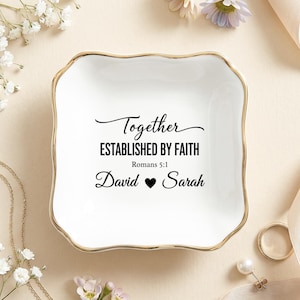 May include: A square white ceramic trinket dish with scalloped edges and a gold-painted rim. The center features black text reading Together ESTABLISHED BY FAITH Romans 5:1 David heart Sarah. The dish has a smooth, glossy finish and curved sides.