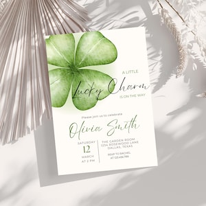 Puede incluir: Una tarjeta de invitación blanca con un trébol verde acuarela y el texto "A Little Lucky Charm Is On The Way". La tarjeta también incluye detalles para un evento, incluyendo el nombre, la fecha, la hora y el lugar.
