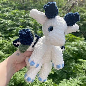 May include: A handmade white crochet plush toy with blue accents. The toy features blue eyes, blue paw pads, and dark blue tufts on its head. The plush has a whimsical design, possibly representing a mythical creature or animal.