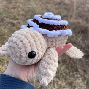 Puede incluir: Un juguete de tortuga de ganchillo con un cuerpo beige suave y un ojo negro redondo. El caparazón está diseñado para parecerse a un rollo de canela, con capas de hilo marrón oscuro cubiertas con espirales de hilo blanco, creando una textura.