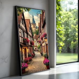 May include: A framed print depicting a picturesque European street. The scene shows a cobbled street lined with half-timbered buildings, red-tiled roofs, and pots of bright pink flowers. A castle with a tall spire is visible in the background, under a blue sky.