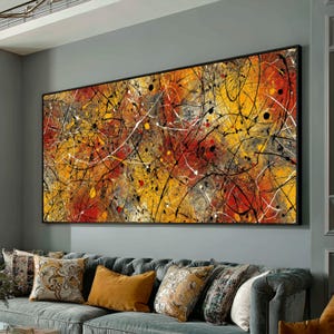 Könnte beinhalten: Abstrakter Kunstdruck auf Leinwand mit schwarzem Rahmen. Das Kunstwerk zeigt eine lebendige Mischung aus Farben, darunter Gelb, Rot und Grau, mit schwarzen und weißen Farbspritzern und Linien. Das Gemälde wird über einem grauen Sofa mit dekorativen Kissen ausgestellt.