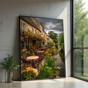 Könnte beinhalten: Gerahmter Kunstdruck eines malerischen Cafés in Europa. Das Bild zeigt ein Steingebäude mit Tischen und Stühlen im Freien, geschmückt mit bunten Blumen. Ein Kanal verläuft neben dem Café, umgeben von üppigem Grün und einem bewölkten Himmel.
