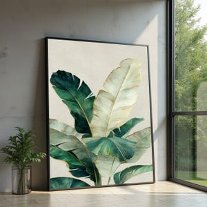 Puede incluir: Una impresión botánica enmarcada con grandes hojas de plátano estilizadas en tonos verdes y crema, acentuadas con detalles dorados. La obra de arte se presenta sobre un fondo neutro, creando una estética moderna y elegante. El marco es negro.