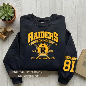 May include: Black sweatshirt with "RAIDERS BOSTON HOCKEY" in gold lettering. Features a shield with an "R" and crossed hockey sticks. The sleeve displays "ROZANOV 81" in gold. The text "EST.1924" is also visible.