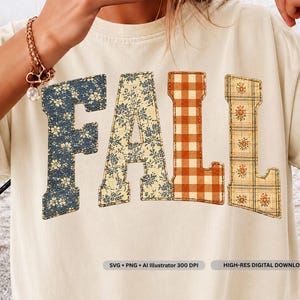 May include: A cream-coloured t-shirt featuring the word FALL in large block letters. Each letter has a distinct pattern: the F is dark blue with small flowers, the A is cream with blue vines, the L is orange gingham, and the L is plaid with flowers.