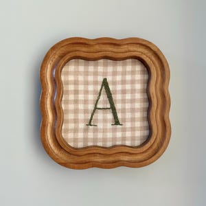 Op de afbeelding: Een vierkante houten lijst met een golvende, geschulpte rand met daarin een stuk lichtbeige en witte gingham-stof. Gecentreerd op de stof staat een enkele hoofdletter A geborduurd in een donker bosgroene draad met een zichtbare steektextuur.