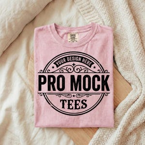 Puede incluir: Una camiseta rosa claro doblada con un dise&ntilde;o circular negro que dice "PRO MOCK TEES" y "YOUR DESIGN HERE". La camiseta est&aacute; sobre una tabla de madera.