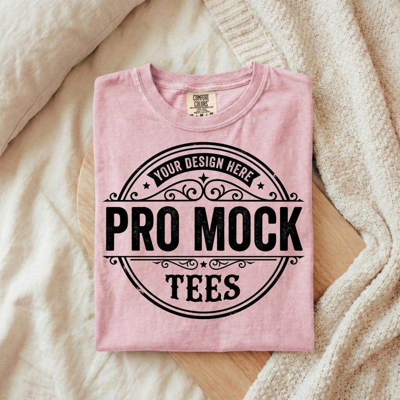 Puede incluir: Una camiseta rosa claro doblada con un dise&ntilde;o circular negro que dice "PRO MOCK TEES" y "YOUR DESIGN HERE". La camiseta est&aacute; sobre una tabla de madera.