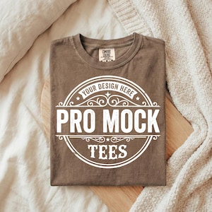 Puede incluir: Una camiseta marr&oacute;n doblada con un gr&aacute;fico blanco que dice "YOUR DESIGN HERE" y "PRO MOCK TEES". La camiseta tiene cuello redondo y una peque&ntilde;a etiqueta en el cuello. La camiseta est&aacute; sobre una superficie de madera.