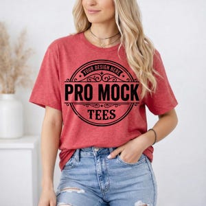 May include: A red t-shirt with the text "PRO MOCK TEES" in a black circular design. The shirt is short-sleeved and made of a soft material. The model is wearing blue jeans and has blonde hair.