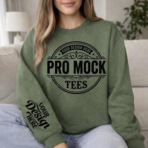 May include: Olive green sweatshirt with the text "PRO MOCK TEES" in a circular design on the front. The sleeve features the text "Your Design Here". The sweatshirt is a mock-up for apparel designs.