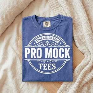 Puede incluir: Una camiseta azul doblada con un gr&aacute;fico blanco que dice "YOUR DESIGN HERE" y "PRO MOCK TEES" en un dise&ntilde;o circular. La camiseta est&aacute; hecha de un material suave y tiene una etiqueta Comfort Colors. La camiseta est&aacute; sobre una superficie de madera.