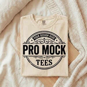 Puede incluir: Una camiseta beige Comfort Colors con un gr&aacute;fico circular negro que dice "Your Design Here" en la parte superior, "Pro Mock" en el centro y "Tees" en la parte inferior. La camiseta est&aacute; doblada sobre una superficie de madera.