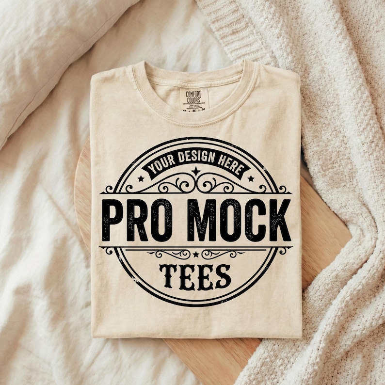 Puede incluir: Una camiseta beige Comfort Colors con un gr&aacute;fico circular negro que dice "Your Design Here" en la parte superior, "Pro Mock" en el centro y "Tees" en la parte inferior. La camiseta est&aacute; doblada sobre una superficie de madera.