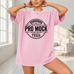 May include: A light pink t-shirt with a large black circular graphic that reads "YOUR DESIGN HERE" at the top, and "PRO MOCK TEES" in the center. The shirt has short sleeves and a relaxed fit.