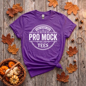 May include: A purple short-sleeve t-shirt featuring a white circular graphic print with the text "YOUR DESIGN HERE", "PRO MOCK", and "TEES" centered on the chest. The shirt has a crew neckline and a tag visible at the inner collar.
