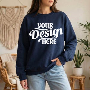 May include: Navy blue sweatshirt with the text "YOUR Design HERE" in white script. The long-sleeved crew neck top is a solid color and has a relaxed fit. The sweatshirt is shown on a person wearing blue jeans.