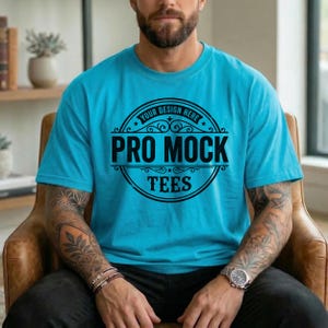 May include: A turquoise blue t-shirt with a crew neck. The shirt features a black circular design with the text "YOUR DESIGN HERE" at the top, and "PRO MOCK TEES" in the center. The person is wearing a watch and has tattoos on their arms.