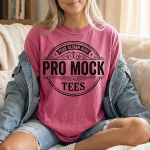 May include: A pink t-shirt with a black circular design that reads "YOUR DESIGN HERE PRO MOCK TEES". The shirt is a solid color and has a relaxed fit. The model is wearing a denim jacket.