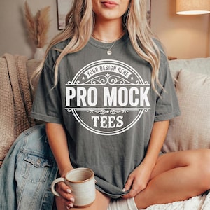 May include: A gray t-shirt with a white circular design that reads "Your Design Here" and "Pro Mock Tees". The shirt is worn by a person sitting down, holding a ceramic mug. A denim jacket is draped over the arm of the couch.