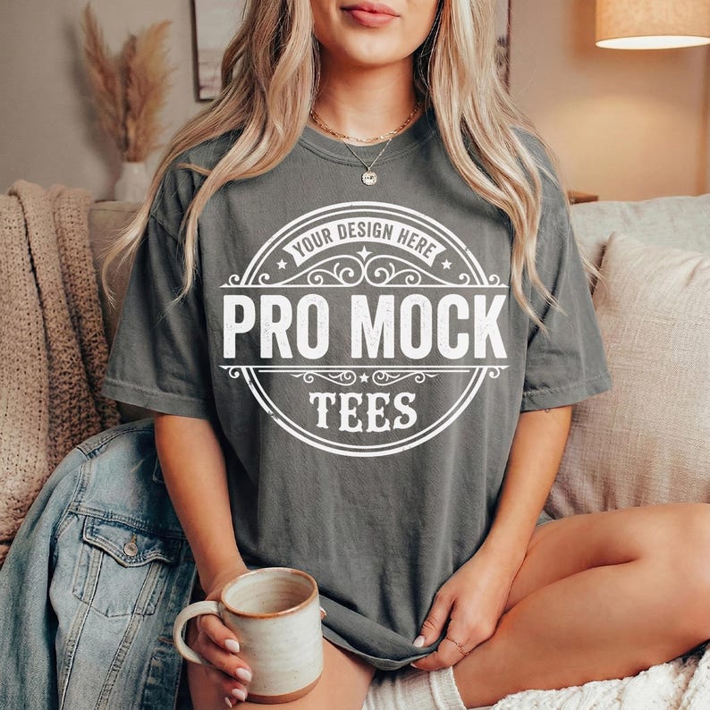 May include: A gray t-shirt with a white circular design that reads "Your Design Here" and "Pro Mock Tees". The shirt is worn by a person sitting down, holding a ceramic mug. A denim jacket is draped over the arm of the couch.