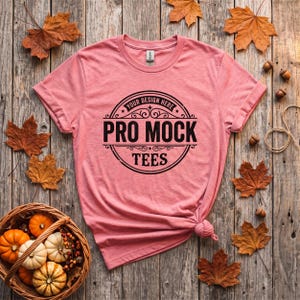 May include: A pink short-sleeve t-shirt featuring a black circular graphic with decorative scrollwork and the text "YOUR DESIGN HERE PRO MOCK TEES" printed in the center. The shirt has a crew neckline and a small tag visible at the inner collar.